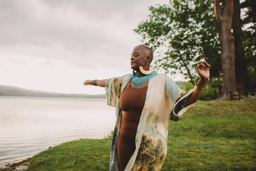 Black woman standing at the edge of a lake with eyes closed and arms outstretched.